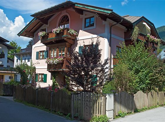 Ein charmantes, pinkes Gebäude mit Balkonen, die mit Blumen geschmückt sind. Darum herum sieht man gepflegte Gärten und eine ruhige Straßenansicht.