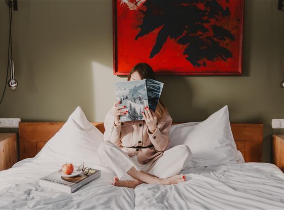 Eine Person sitzt auf einem Bett und hält ein Magazin hoch. Im Hintergrund sind ansprechende Wandkunst und eine gemütliche Zimmeratmosphäre sichtbar.