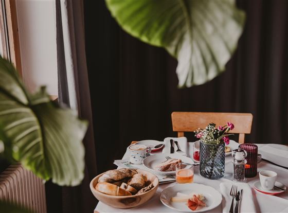 Ein schön gedeckter Tisch mit frischem Frühstück. Es gibt Brot, Obst und ein Getränk, umgeben von grünen Pflanzen.