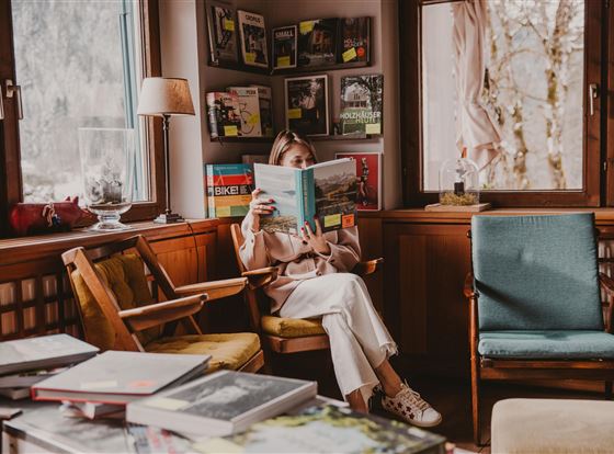 Eine Frau sitzt in einem gemütlichen Raum und liest ein Buch. Im Hintergrund sind Regale mit verschiedenen Zeitschriften und Büchern zu sehen.