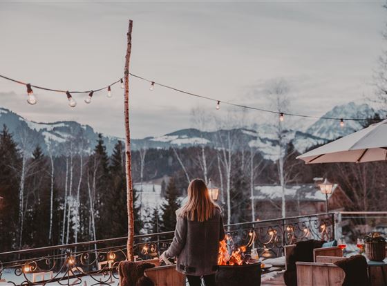 Eine Frau steht vor einem Feuerscheit auf einer Terrasse mit Blick auf die Berge. Lichterketten und eine entspannte Atmosphäre schaffen eine gemütliche Stimmung.