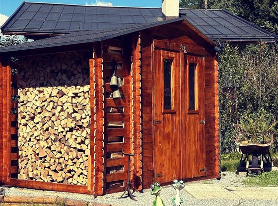 Eine gemütliche Holzscheune mit einer Wand aus aufgestapeltem Brennholz. Im Vordergrund stehen zwei dekorative Figuren in Form von Fröschen.