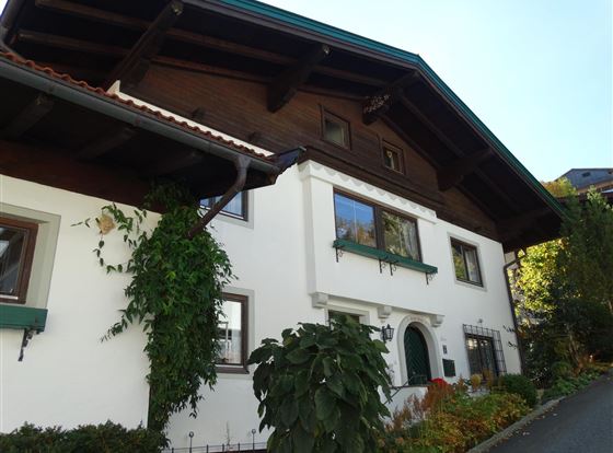 Ein traditionelles Haus mit einem Holzdach und grünen Fensterrahmen. Vor dem Haus sind Pflanzen und ein gepflegter Garten zu sehen.
