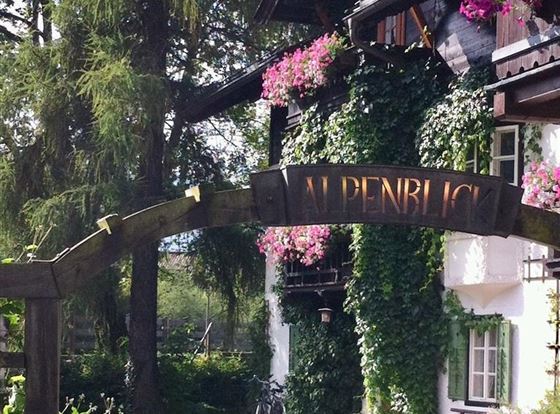 Ein charmantes Haus mit einem begrünten Eingangsschild „Alpenblick“. Die Fassade ist von bunten Blumen und Pflanzen umgeben.