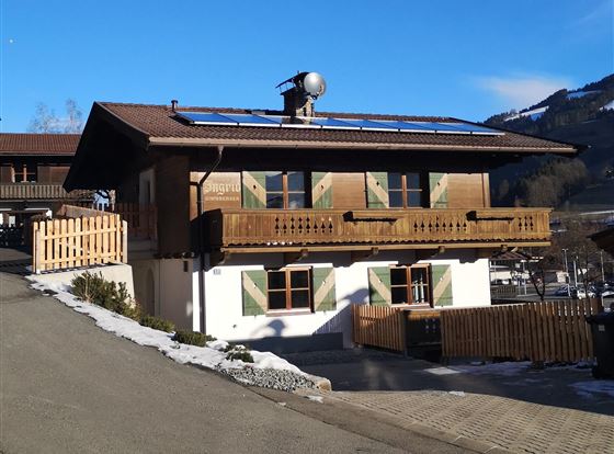 Ein traditionelles alpenländisches Haus mit einem Holzbalcony und grünen Fensterläden. Die Umgebung ist schneebedeckt und die Berge sind im Hintergrund sichtbar.