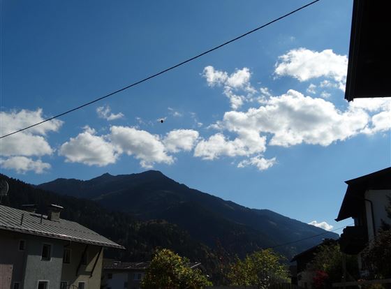 Ein klarer Himmel mit einigen Wolken über den Bergen. Im Vordergrund sind Häuser und Bäume zu sehen.