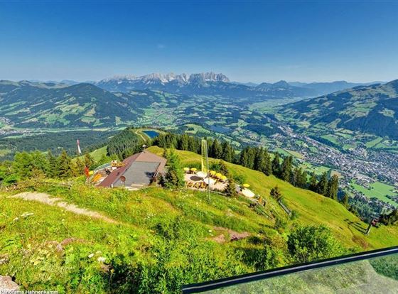 Eine malerische Berglandschaft mit grünen Wiesen und einem klaren Himmel. Im Vordergrund ist eine Alm mit Sitzgelegenheiten zu sehen, während die Berge im Hintergrund majestätisch hervortreten.