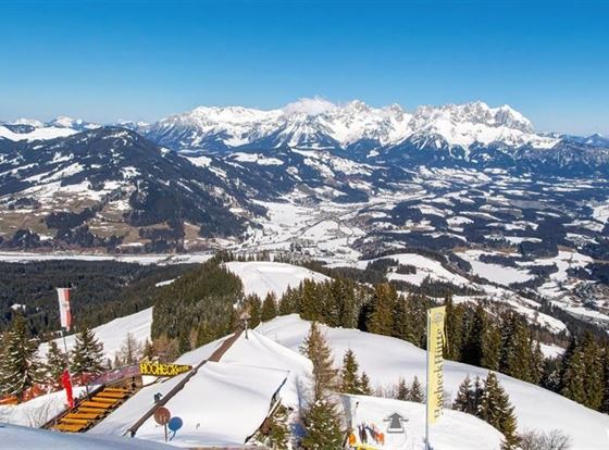 Eine atemberaubende Berglandschaft mit schneebedeckten Gipfeln und einem klaren blauen Himmel. Im Vordergrund sind verschneite Pisten und Bäume zu sehen.