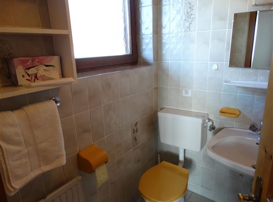 Ein kleines Badezimmer mit hellen Fliesen. Es enthält eine Toilette mit einem gelben Deckel und ein Waschbecken.