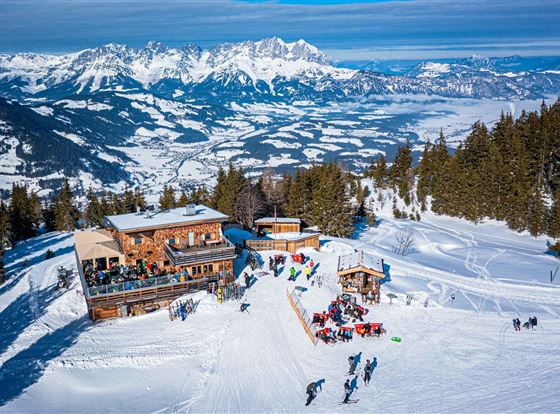 Eine malerische Berglandschaft mit schneebedeckten Pisten und einer gemütlichen Hütte. Im Hintergrund erstrecken sich majestätische Berge unter einem klaren blauen Himmel.