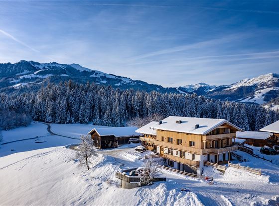 Ein malerisches Bergpanorama mit schneebedeckten Hügeln und einem traditionellen Holzhaus. Die Umgebung ist idyllisch und von Nadelbäumen umgeben.