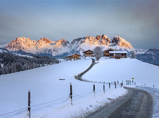 Eine schneebedeckte Landschaft mit malerischen Bergen im Hintergrund. Kleine Holzhäuser und ein verschneiter Weg führen durch die winterliche Szene.