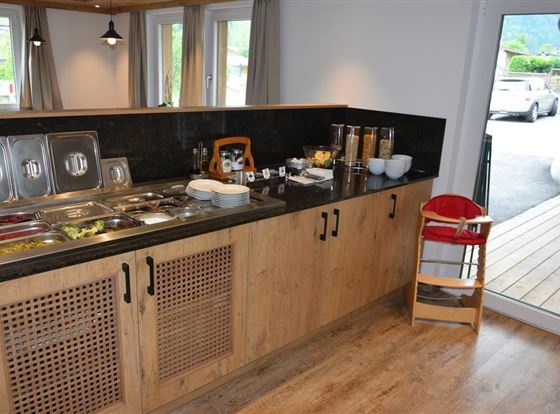 Ein moderner Frühstücksbereich mit einem Holzschrank und verschiedenen Zutaten in Schalen. Im Hintergrund ist ein Stuhl für Kleinkinder und ein Fenster mit Blick nach draußen zu sehen.