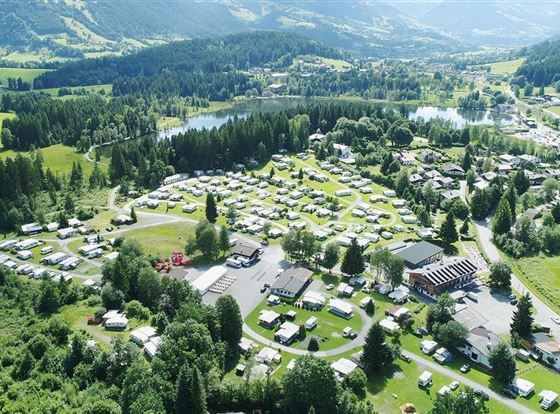 Ein großer Campingplatz in einer grünen Umgebung mit vielen Wohnmobilen und Stellplätzen. Im Hintergrund sieht man sanfte Hügel und einen kleinen See.
