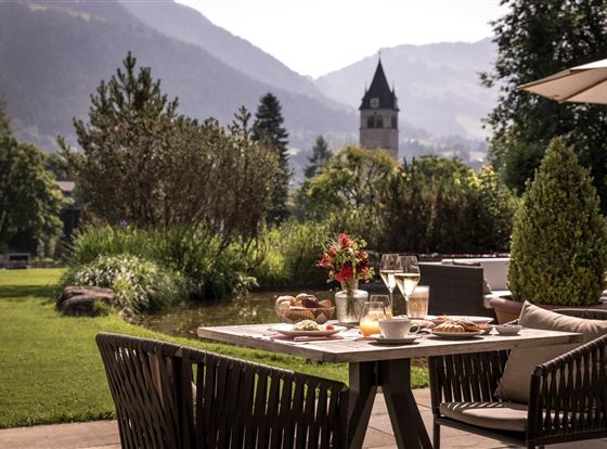 Ein schöner Garten mit einem Tisch, auf dem Frühstück und Getränke serviert werden. Im Hintergrund sind Berge und ein Kirchturm sichtbar.