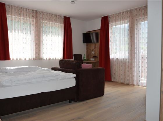 Ein helles Zimmer mit einem Doppelbett und einem gemütlichen Sessel. Große Fenster lassen viel Licht hinein und die roten Vorhänge sorgen für einen warmen Akzent.