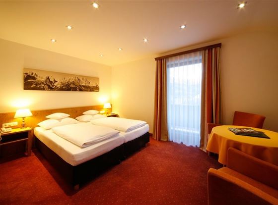 Ein gemütliches Hotelzimmer mit einem Doppelbett und warmen Farben. Es gibt ein Fenster mit Vorhängen und einen kleinen Tisch mit Stühlen.