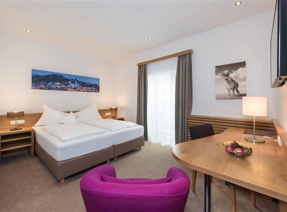 Ein modernes Hotelzimmer mit zwei Betten und einem Schreibtisch. Es gibt eine helle Atmosphäre mit einer großen Fensterfront und farbenfrohen Akzenten.