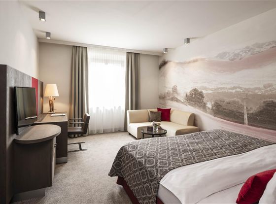 A modern hotel room with a large bed and stylish decor. There is a TV, a desk, and plenty of natural light from the window.