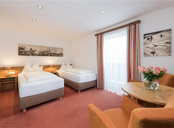 Ein gemütliches Hotelzimmer mit zwei Betten und einem Fenster, das viel Tageslicht bietet. Ein runder Tisch mit einem Blumenstrauß und eine gemütliche Sitzecke runden das Ambiente ab.