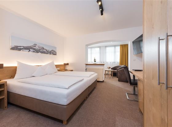 Ein modernes Hotelzimmer mit zwei Betten, einem Schreibtisch und einer gemütlichen Sitzecke. Große Fenster sorgen für viel Tageslicht.