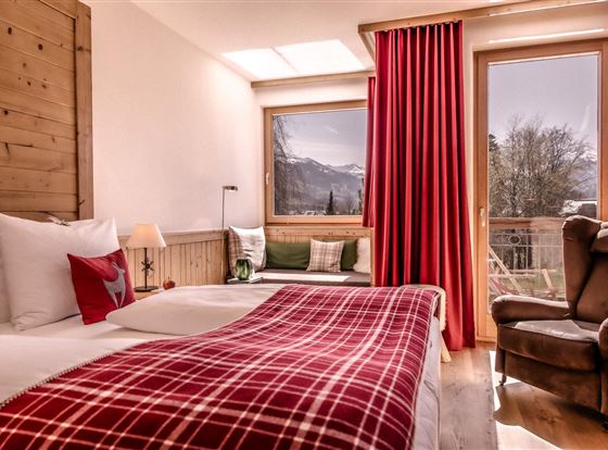 Ein gemütliches Hotelzimmer mit einem Doppelbett und einer rot karierten Decke. Durch das Fenster hat man einen Blick auf die Berge und viel Tageslicht.