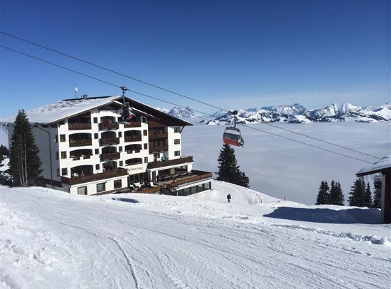 Ein schneebedecktes Bergpanorama mit einem gemütlichen Hotel und einer Seilbahn. Die Aussicht zeigt die beeindruckenden Berge und einen wolkenverhangenen Himmel.