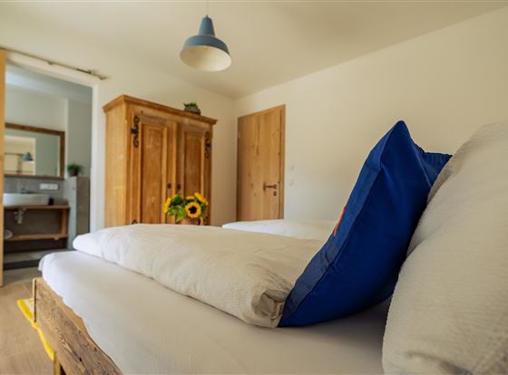 Ein gemütliches Schlafzimmer mit zwei Betten und einem Holzschrank. Im Hintergrund sieht man ein Badezimmer mit Spiegel und kleiner Deko.