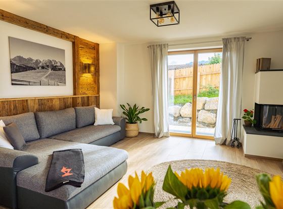 Ein modernes Wohnzimmer mit einer gemütlichen grauen Couch und großen Fenstern. Der Raum ist hell und freundlich, mit einem schönen Blick auf den Außenbereich.