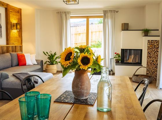 Ein gemütlicher Wohnraum mit einem Essbereich und einer modernen Couch. Auf dem Tisch steht eine Vase mit Sonnenblumen und Glasflaschen.