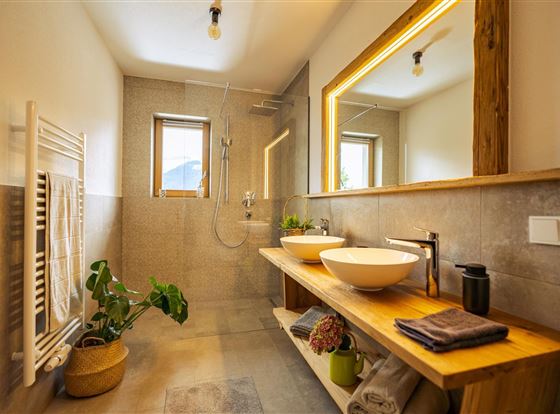 Ein modernes Badezimmer mit natürlichen Holzdetails. Es verfügt über zwei Waschbecken, eine große Dusche und einen kleinen Pflanzenakzent.