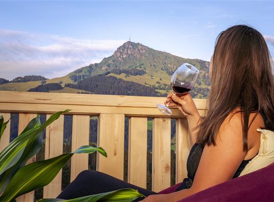 Eine Frau sitzt auf einem Balkon und genießt ein Glas Rotwein. Im Hintergrund ist ein malerischer Berg mit grünen Wiesen zu sehen.