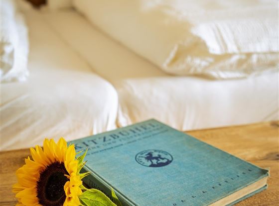 Ein gemütliches Schlafzimmer mit zwei Betten und frischen weißen Bettwäsche. Auf einem Holztisch liegt ein blaues Buch und eine Sonnenblume.