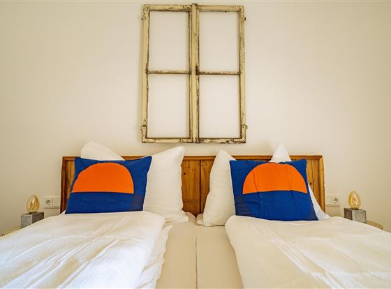 Ein gemütliches Schlafzimmer mit zwei Betten und bunten Kissen. An der Wand hängt ein altes Fenster als Dekoration.