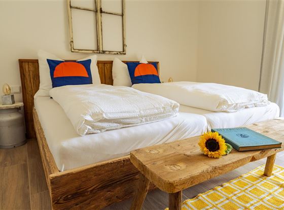 Ein gemütliches Schlafzimmer mit zwei Holzbetten und bunten Kissen. Auf dem Tisch liegt ein Buch und eine Sonnenblume.
