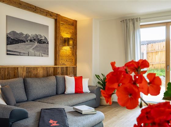 Ein gemütlicher Wohnbereich mit einem grauen Sofa und roten Kissen. Im Hintergrund sind eine Wand mit Holzverkleidung und ein großes Fenster zu sehen, durch das viel Licht fällt.