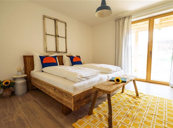 Ein helles Schlafzimmer mit einem bequemen Doppelbett und bunten Kissen. Große Fenster lassen viel Licht hinein und es gibt eine gemütliche Tisch mit einer Sonnenblume.