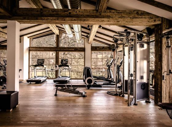 Ein moderner Fitnessraum mit Holzverkleidung und großen Fenstern. Es sind verschiedene Fitnessgeräte wie Laufbänder und Hantelstationen vorhanden.