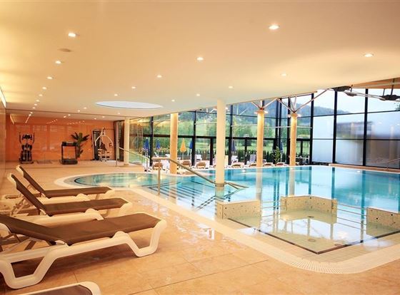 Ein elegantes Hallenbad mit bequemen Liegen und großen Fenstern. Das Wasser glitzert und der Raum ist hell und einladend.