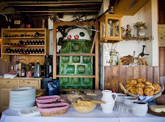Eine gemütliche Stube mit einem großen Ofen im Hintergrund. Auf dem Tisch sind verschiedene Speisen, Geschirr und Getränke angerichtet.