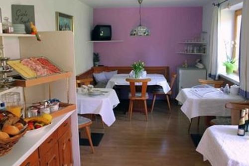 Ein einladender Frühstücksraum mit weißen Tischen und Stühlen. Auf einem Buffet sind frische Brötchen und Obst bereitgestellt.