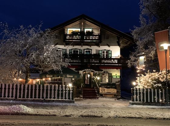 Ein gemütliches Haus im winterlichen Schnee, beleuchtet von warmen Lichtern. Die Umgebung ist von schneebedeckten Bäumen und einer frostigen Atmosphäre geprägt.