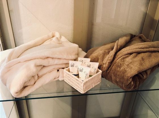 Zwei kuschelige Decken in hellen Farben liegen auf einem Glasregal. Darunter befindet sich ein kleiner Korb mit mehreren Pflegeprodukten.