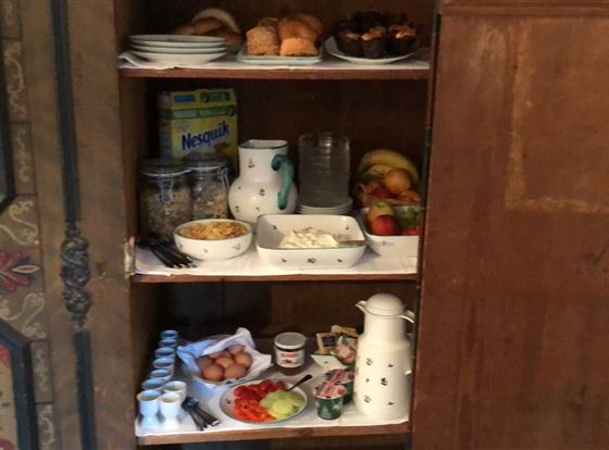 Ein rustikaler Schrank mit mehreren Regalen voller Lebensmittel. Es gibt Obst, Gebäck, Joghurt und Getränke auf den Regalen.