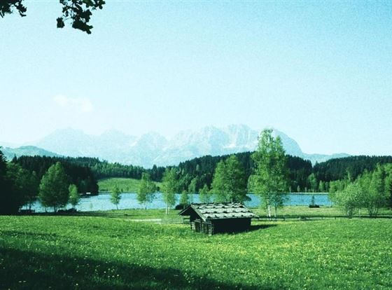 Eine malerische Landschaft mit einem ruhigen See und Bergen im Hintergrund. Auf der Wiese befindet sich eine kleine, rustikale Hütte.