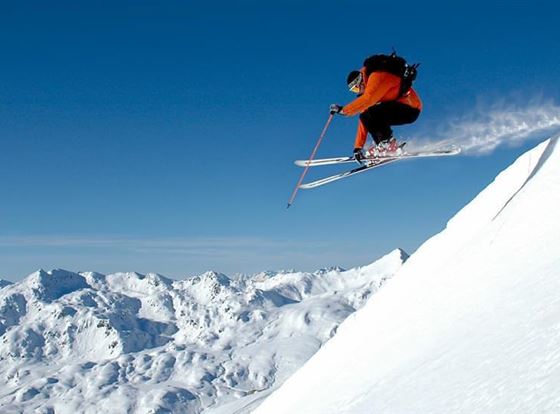 Ein Skifahrer springt von einem schneebedeckten Gipfel in die Luft. Im Hintergrund sind majestätische Berge und ein klarer blauer Himmel sichtbar.