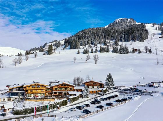 Eine malerische Winterlandschaft mit schneebedeckten Hügeln und einer klaren blauen Himmel. Im Vordergrund befinden sich gemütliche Chalets und ein Parkplatz.