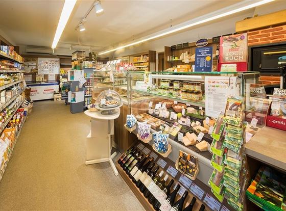 Ein gut sortierter Laden mit Regalen voller Lebensmittel und frischen Produkten. Es gibt eine Auslage mit Käse und anderen Delikatessen.