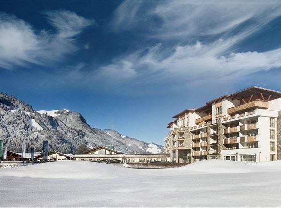Ein modernes Hotelgebäude in einer verschneiten Landschaft. Im Hintergrund sind majestätische Berge und ein klarer blauer Himmel zu sehen.