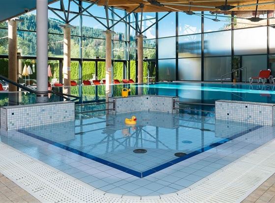 Ein modernes Hallenbad mit hellem Wasser und großen Fenstern. In der Mitte schwimmt ein gelbes Gummienten-Spielzeug.